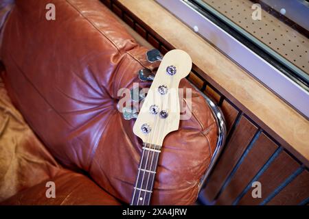 Eine E-Bassgitarre liegt horizontal über einem abgenutzten braunen Ledersofa, dessen Kopfstock vor einem verschwommenen Innenhintergrund fokussiert ist. Stockfoto