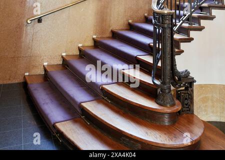 Eine polierte Wendeltreppe aus Holz mit violettem Teppichboden, einem Handlauf aus Metall und einem kunstvollen Geländer. Stockfoto