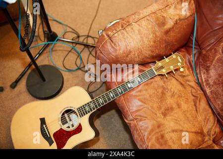 Eine Akustikgitarre liegt auf einer braunen Ledercouch mit Kabeln auf dem Boden und einem Mikrofonständer im Hintergrund. Stockfoto
