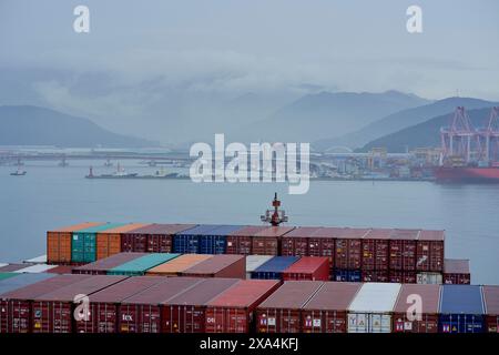 Stapel von bunten Schiffscontainern in einem Hafen mit Kränen und Schiffen im Hintergrund, unter einem bedeckten Himmel mit Bergen in der Ferne. Stockfoto