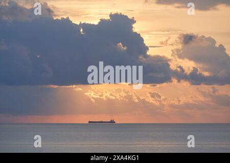 Eine ruhige Ozeanszene in der Abenddämmerung mit einem Frachtschiff, das am Horizont sichtbar ist, beleuchtet von den sanften, warmen Farbtönen der untergehenden Sonne, die durch verstreute Wolken schaut. Stockfoto