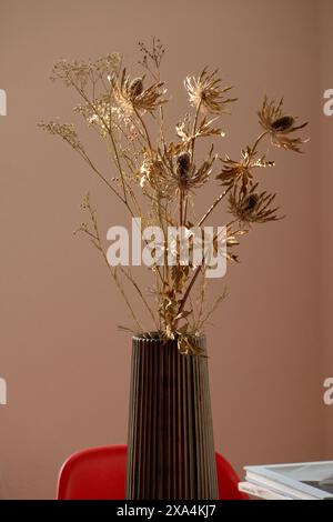 Ein Strauß getrockneter Blumen ist in einer gerippten Vase auf einem Tisch vor dem Hintergrund einer pfirsichfarbenen Wand und eines roten Stuhls angeordnet. Stockfoto