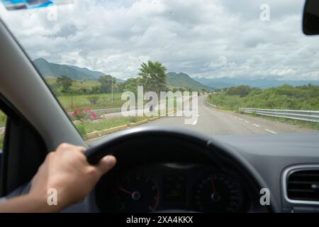 Ein Blick aus der Perspektive des Fahrers in ein Fahrzeug, mit der linken Hand des Fahrers am Lenkrad, mit Blick auf eine kurvenreiche Straße, die von grünen Landschaften unter bewölktem Himmel flankiert wird. Stockfoto