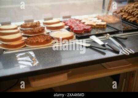 Eine Auswahl an Kuchen und Gebäck, die in einer Bäckerei hinter einer Glastheke präsentiert werden, mit Servierutensilien im Vordergrund. Stockfoto