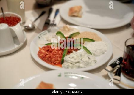 Eine Auswahl griechischer Dips auf einem weißen Teller mit Gurkenscheiben und schwarzer Olivengarnitur sowie ein alkoholfreies Getränk und ein Tisch. Stockfoto