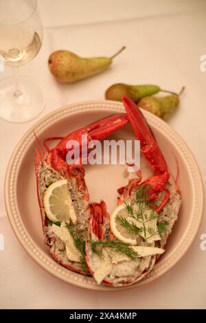 Ein überzogenes Meeresfrüchte-Gericht besteht aus einem gekochten Hummer, garniert mit Zitrone und Dill, begleitet von einem Glas Weißwein und zwei Birnen im Hintergrund, alles auf einem eleganten Tisch. Stockfoto