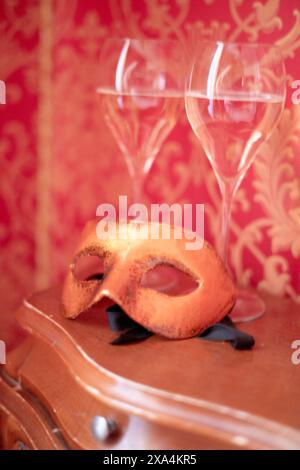 Eine orangene Maske liegt auf einem antiken Holztisch vor einem roten Hintergrund mit Tapeten und zwei leeren Weingläsern dahinter. Stockfoto