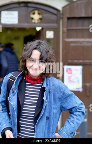 Vor einer Holztür steht eine lächelnde Frau mit lockigem Haar, einer Jeansjacke, einem gestreiften Hemd und einem roten Schal. Stockfoto