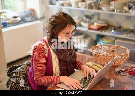 Eine Frau sitzt an einem Tisch und arbeitet intensiv an ihrem Laptop in einem Raum mit Küchenregalen im Hintergrund. Stockfoto