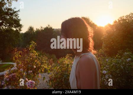 Eine Frau steht in einem Garten bei Sonnenuntergang, das warme Licht strahlt ein weiches Licht auf ihr Profil mit blühenden Blumen und Grün im Hintergrund. Stockfoto