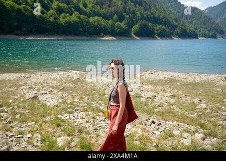 Eine Frau in rotem Kleid spaziert entlang eines felsigen Ufers an einem malerischen See, umgeben von bewaldeten Hügeln unter blauem Himmel. Stockfoto