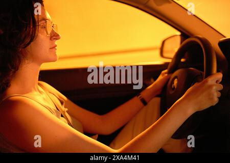 Eine Frau sitzt auf dem Fahrersitz eines Autos, Hände am Lenkrad, mit einem fokussierten Blick auf ihrem Gesicht, beleuchtet von einem warmen, orangen Licht. Stockfoto