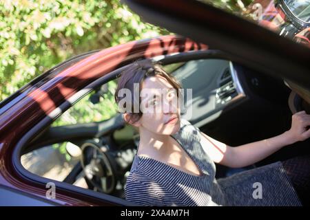 Eine Frau sitzt in einem Auto, blickt durch ein geöffnetes Autofenster, mit Bäumen und Sonnenlicht im Hintergrund, die Schatten über ihr Gesicht werfen. Stockfoto