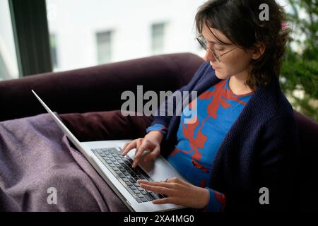 Eine Frau sitzt auf einer violetten Couch mit einem Laptop. Sie scheint sich auf ihre Arbeit konzentriert zu haben, wobei natürliches Licht den Raum von einem Fenster neben ihr beleuchtet. Stockfoto