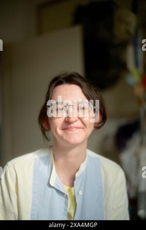 Eine lächelnde junge Frau mit kurzen Haaren, die eine runde Brille und ein hellblaues Hemd über einem gelben Oberteil trägt, steht drinnen, mit einem verschwommenen Hintergrund, der auf einen Raum mit Gegenständen hindeutet. Stockfoto