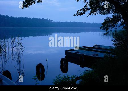 Eine ruhige Seeszene am frühen Morgen mit Ruderbooten, die an einem Dock gebunden sind, mit einer ruhigen, reflektierenden Wasseroberfläche und einem Hauch von Nebel, der über dem See schwebt. Stockfoto