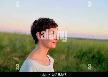 Eine junge Frau mit einem kurzen Haarschnitt steht auf einem Feld und blickt in die Ferne während eines ruhigen Sonnenuntergangs. Stockfoto