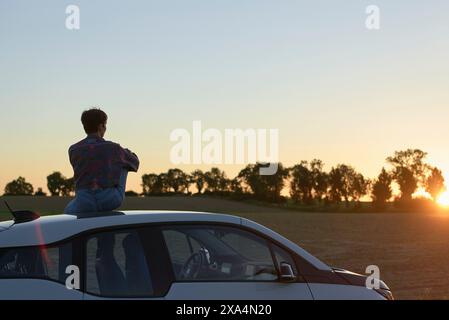 Eine junge Frau sitzt auf einem Auto und schaut den Sonnenuntergang in einer Landschaft. Stockfoto