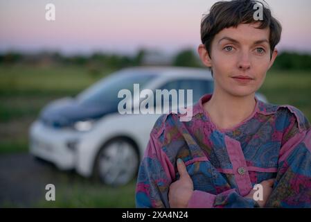 Eine junge Frau mit kurzen Haaren, die ein gemustertes Hemd trägt, steht im Vordergrund mit einem weißen Auto, das in der Abenddämmerung im Hintergrund verschwimmt. Stockfoto