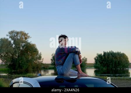 Eine junge Frau sitzt auf dem Dach eines Autos, gewickelt in eine Decke, und blickt in die Ferne mit einem ruhigen Hintergrund eines ruhigen Flusses und einem klaren Himmel in der Abenddämmerung. Stockfoto
