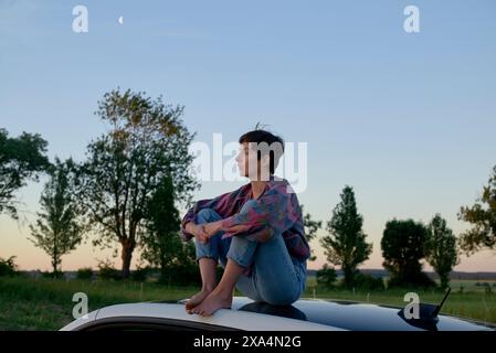 Eine junge Frau mit einem kurzen Haarschnitt sitzt auf einem Auto, in einen bunten Schal gewickelt, vor dem Hintergrund eines offenen Feldes in der Abenddämmerung, mit einem Mondsplitter am Himmel. Stockfoto
