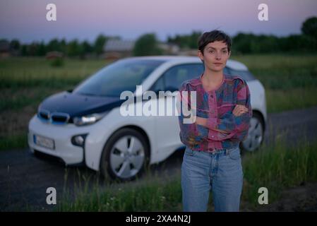 Eine junge Frau steht in der Abenddämmerung selbstbewusst vor einem Elektroauto auf einer Landstraße mit überkreuzten Armen. Stockfoto