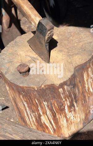 Die Axt ist in ein Holzdeck gehämmert. Als Nächstes ist ein Hammerbock zum Nieten einer Sense. Stockfoto