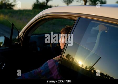 Eine junge Frau sitzt auf dem Fahrersitz eines Autos, deren Silhouette durch das Fenster sichtbar ist, während die Sonne im Hintergrund untergeht und ein warmes Leuchten ausstrahlt. Stockfoto