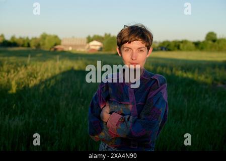 Eine kurzhaarige junge Frau steht auf einem Feld mit überkreuzten Armen und trägt ein lila gemustertes Hemd. Dahinter befindet sich eine üppige grüne Landschaft und ein Haus zur Zeit der goldenen Stunde. Stockfoto