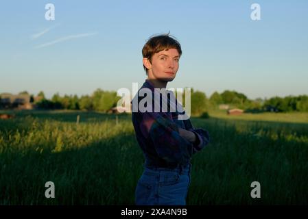 Eine junge Frau steht während der goldenen Stunde auf einem Feld, mit überkreuzten Armen und einem leichten Lächeln, mit kariertem Hemd und Jeans. Stockfoto