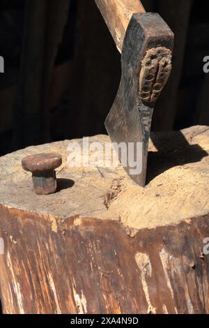 Die Axt ist in ein Holzdeck gehämmert. Als Nächstes ist ein Hammerbock zum Nieten einer Sense. Stockfoto