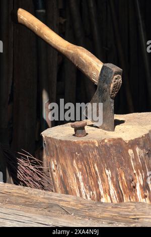 Die Axt ist in ein Holzdeck gehämmert. Als Nächstes ist ein Hammerbock zum Nieten einer Sense. Stockfoto