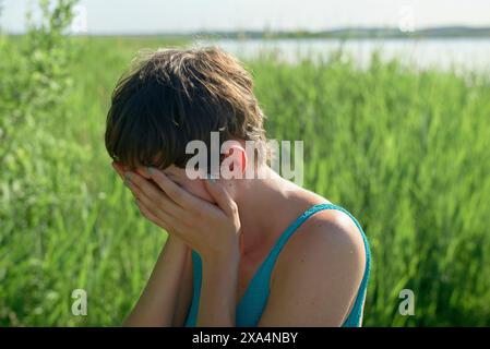 Eine junge Frau sitzt draußen mit dem Gesicht in den Händen und erscheint beunruhigt oder besinnlich im Grünen. Stockfoto