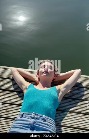 Eine junge Frau liegt auf einer Holzterrasse in der Nähe eines Wassers, sonnt sich im Sonnenlicht mit geschlossenen Augen und Händen hinter dem Kopf. Stockfoto