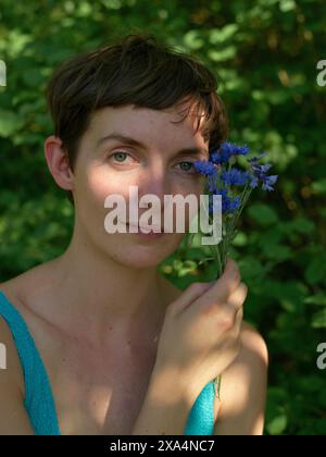 Eine junge Frau mit kurzen Haaren hält einen Blumenstrauß aus blauen Blumen nahe am Gesicht und posiert vor einem grünen natürlichen Hintergrund. Stockfoto