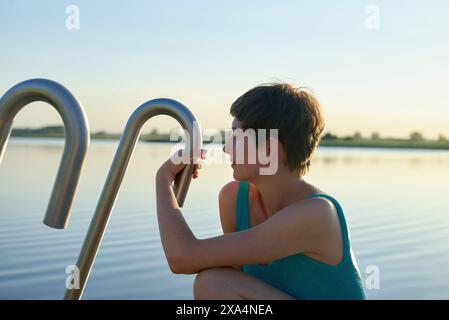 Eine kurzhaarige junge Frau legt ihr Kinn auf ihre Hand, während sie bei Sonnenuntergang an einem ruhigen Wasserkörper sitzt und nachdenklich in die Ferne blickt. Stockfoto