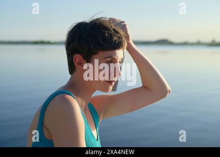 Eine junge Frau mit kurzen Haaren steht in der Abenddämmerung an einem Wasserkörper und hebt ihre Hand an den Kopf, mit einem nachdenklichen Ausdruck und dem Horizont im Hintergrund. Stockfoto