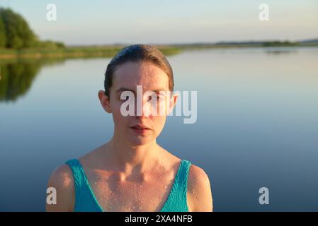 Eine Nahaufnahme einer Frau mit nasser Haut und Haaren, die ein blaues Tanktop trägt und bei Tageslicht vor einem ruhigen Wasserkörper steht. Stockfoto