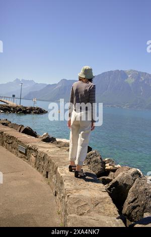 Eine Person steht auf einem felsigen Pfad am See und blickt auf eine Berglandschaft unter einem klaren blauen Himmel. Stockfoto