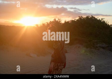 Eine junge Frau steht bei Sonnenuntergang an einem Sandstrand, die Sonne strahlt ein warmes Leuchten und Lichtstrahlen um sie herum. Stockfoto