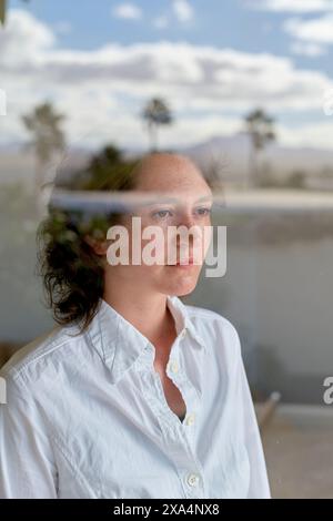 Eine nachdenkliche junge Frau mit lockigem Haar blickt aus einem Fenster, ihre Reflexion und ferne Bäume mischen sich im Bild. Stockfoto