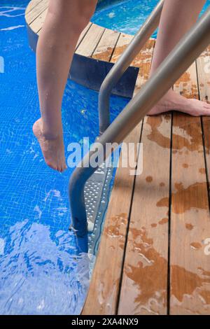 Eine junge Frau taucht ihre Zehen von einer Holzterrasse in einen Swimmingpool und hält sich an einer Metallleiter fest. Stockfoto