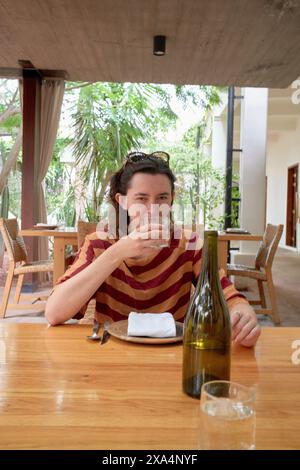 Eine junge Frau, die an einem Esstisch im Freien sitzt, Wasser aus einem klaren Glas trinkt, mit einer leeren Weinflasche im Vordergrund und üppigem Grün im Hintergrund. Stockfoto