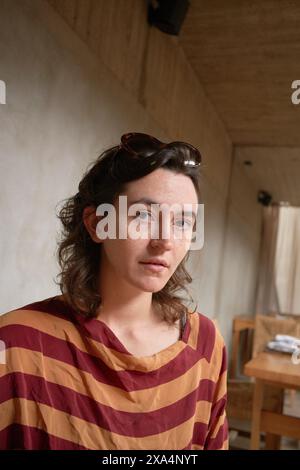 Eine junge Frau mit einer Sonnenbrille auf dem Kopf trägt ein gestreiftes Hemd und blickt in einem Raum mit sanfter Beleuchtung neutral in die Kamera. Stockfoto