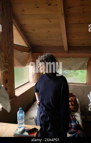 Eine junge Frau steht in einer gemütlichen Holzhütte und blickt aus einem Fenster, in natürliches Licht getaucht. Stockfoto