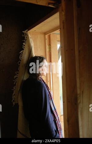 Eine junge Frau steht in einem Holzgestell, das in eine Decke gehüllt ist, und blickt mit nachdenklichem Ausdruck durch ein Fenster. Stockfoto