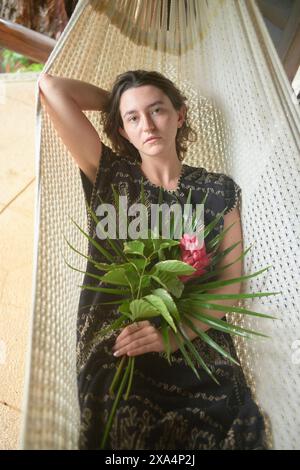 Eine junge Frau sitzt in einer weißen Hängematte mit einem Blumenstrauß aus grünen Blättern und einer roten Blume, gekleidet in einem schwarzen Spitzenkleid, mit einem entspannten und besinnlichen Ausdruck. Stockfoto
