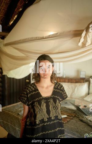 Eine junge Frau steht drinnen mit einem neutralen Ausdruck und trägt ein schwarzes Oberteil mit gemusterten Details. Im Hintergrund befindet sich ein elegantes, drapiertes Baldachin. Stockfoto