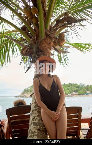 Eine Frau in schwarzem Badeanzug und Sonnenhut steht neben einer Palme an einem Sandstrand mit Meer und Küstenlandschaft im Hintergrund. Stockfoto