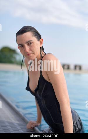 Eine Frau in einem nassen schwarzen Badeanzug taucht aus einem Pool auf, mit Wassertropfen auf der Haut und einem fokussierten Ausdruck. Stockfoto